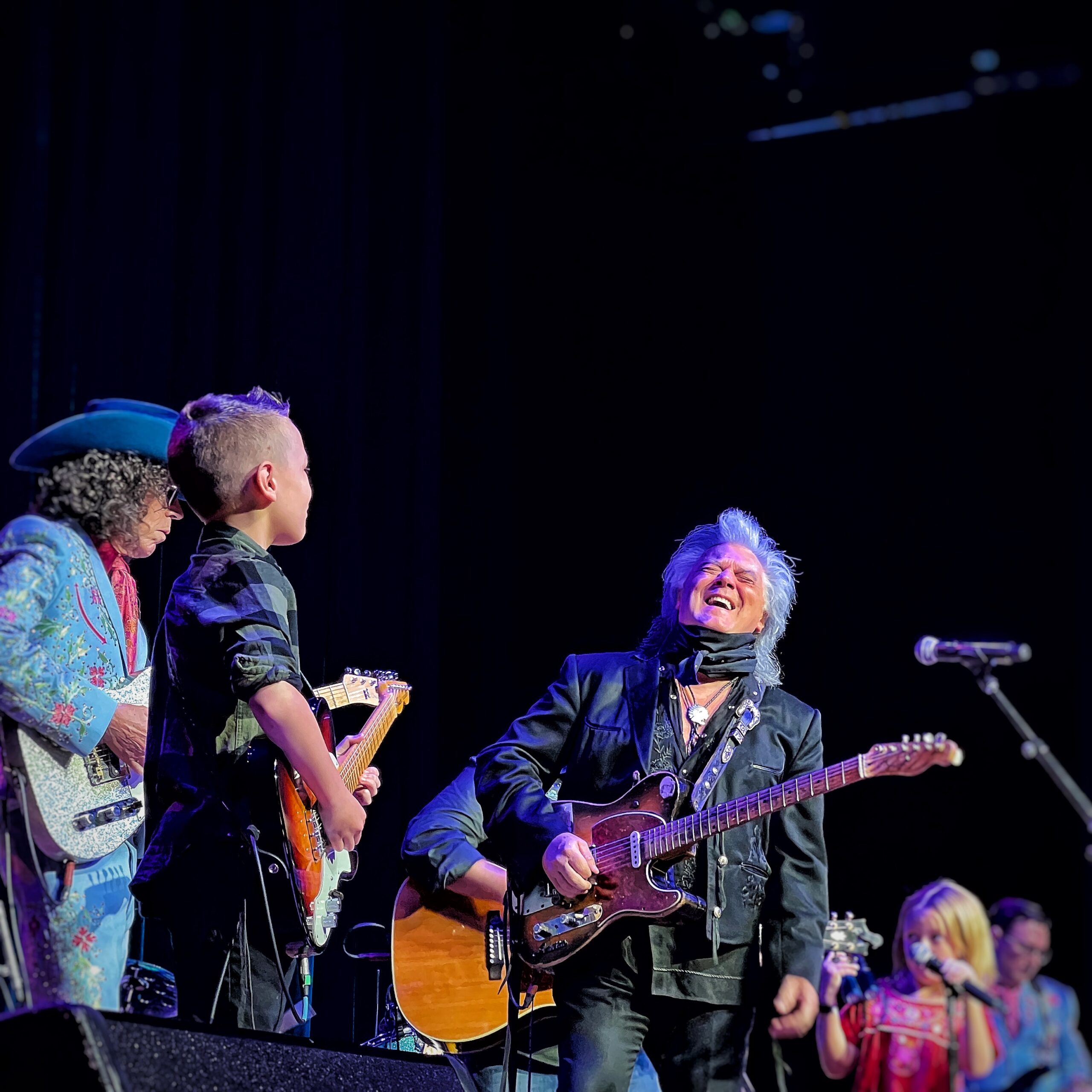 Marty Stuart leaning back laughing next to kid with guitar on stage