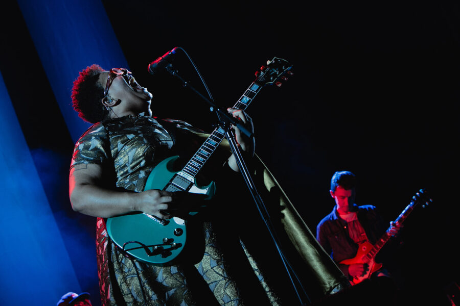 Woman playing guitar and singing into microphone with man on guitar in the background and blue and red lighting