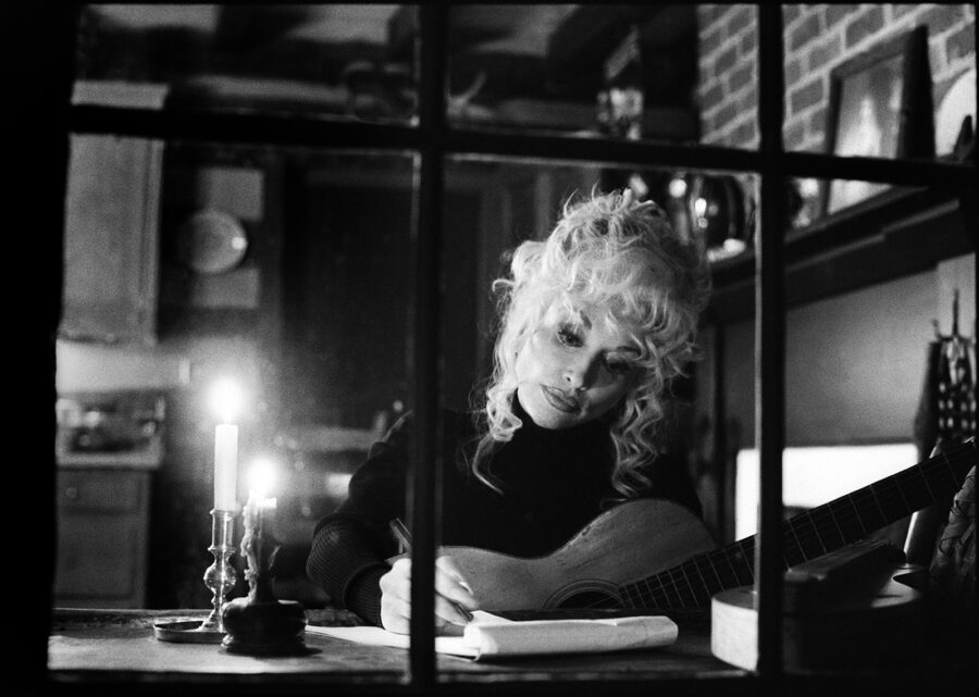 Dolly Parton sitting at her desk holding her guitar writing on a piece of paper.