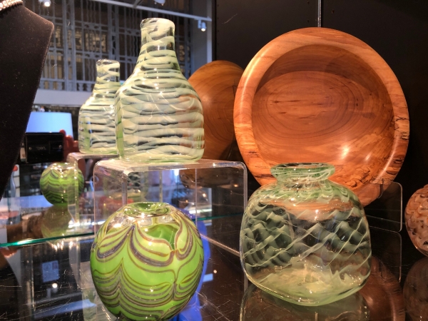 Beautiful glass vases in shades of green are situated alongside a turned wood bowl displayed on a stand so the wood grain is visible.