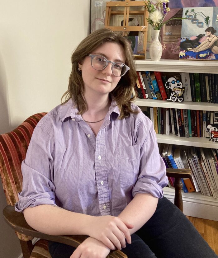 A woman with brown hair sits in a chair in front of a bookshelf.