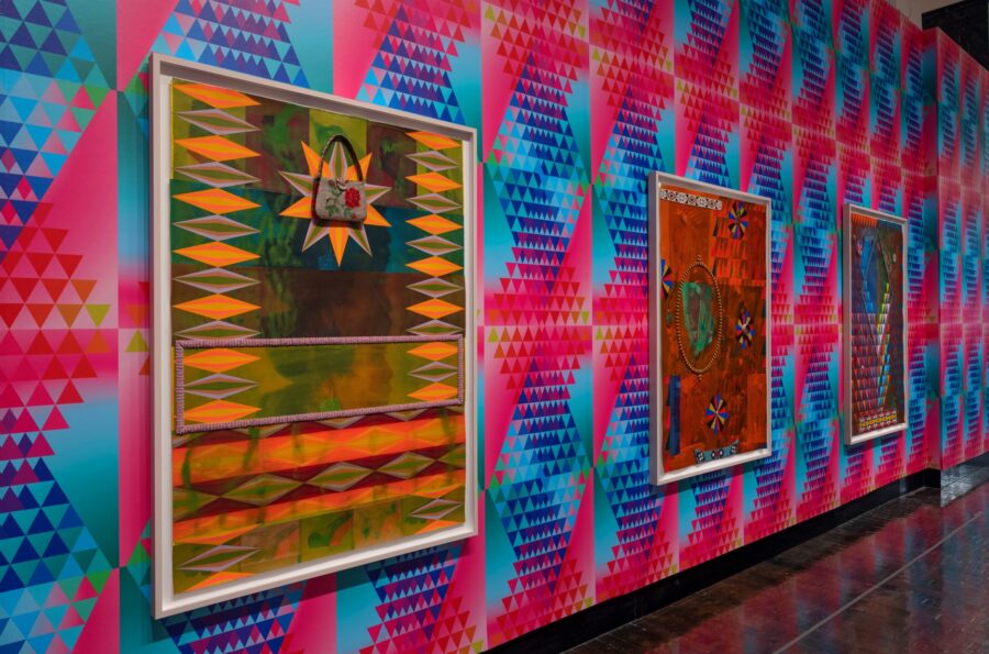 Three large vibrant paintings on a wall covered with pink, blue and green geometric patterns.