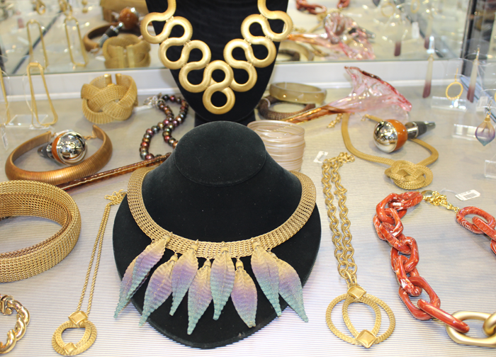 Necklaces, one gold serpentine and the other resembling large feathers are featured on black velvet stands in the center of a case full of necklaces.