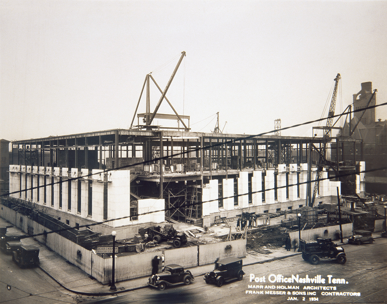 Girders rise and granite cladding appears as construction of the city’s main post office in 1934. Cranes surround the building as construction workers, trucks, and automobile travel around the site.