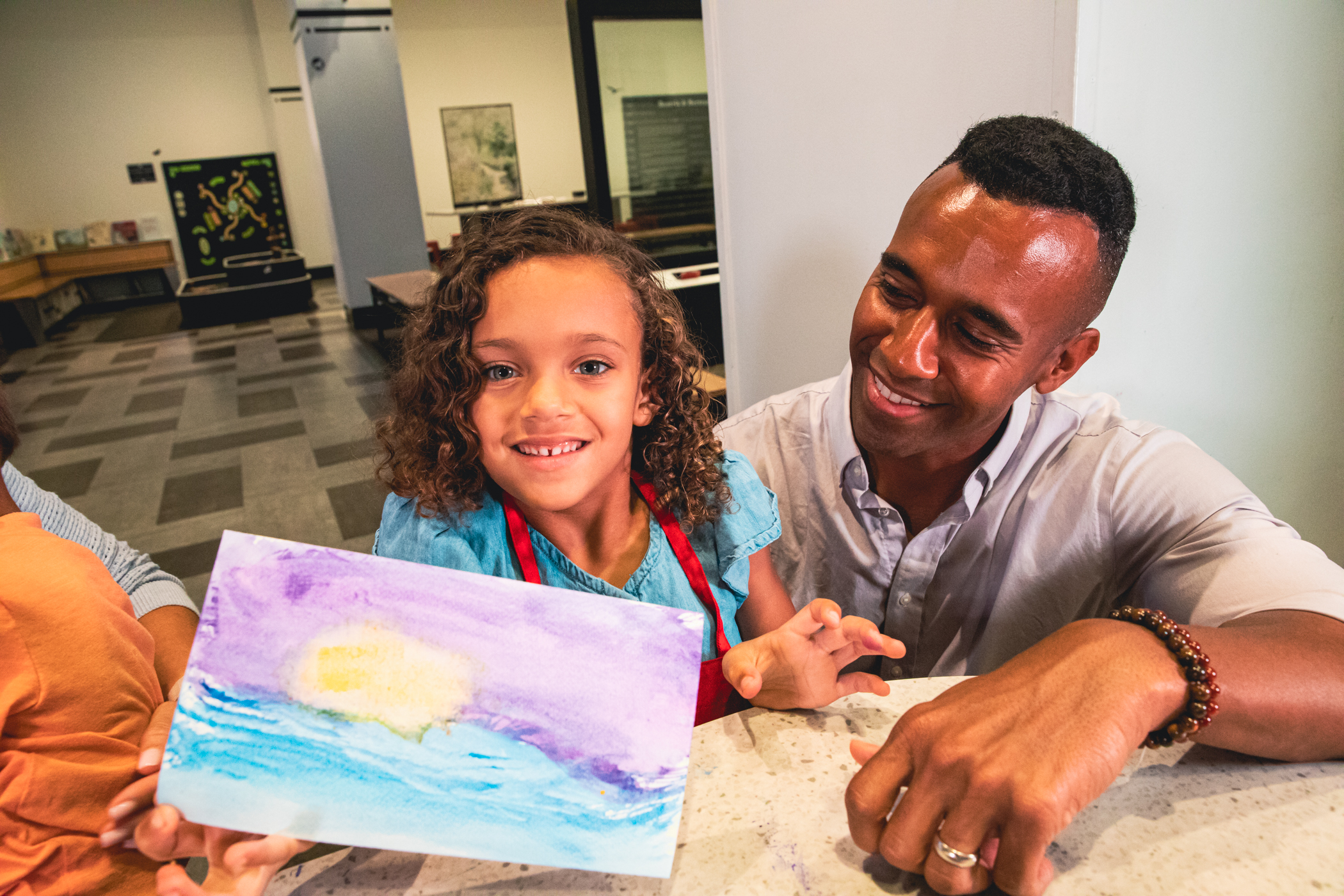 Young girl holding up a painting she made as her dad looks at her and smiles