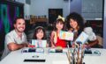 Mom, dad and two young daughters smile and hold up paintings they just made