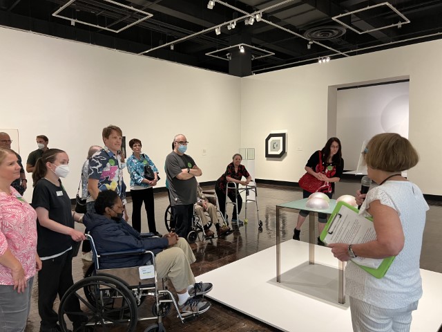 Group of people, some in wheelchairs, listening to a docent give an exhibition tour