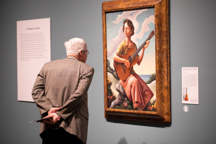 Older man peering close at a painting of a woman holding a guitar outdoors