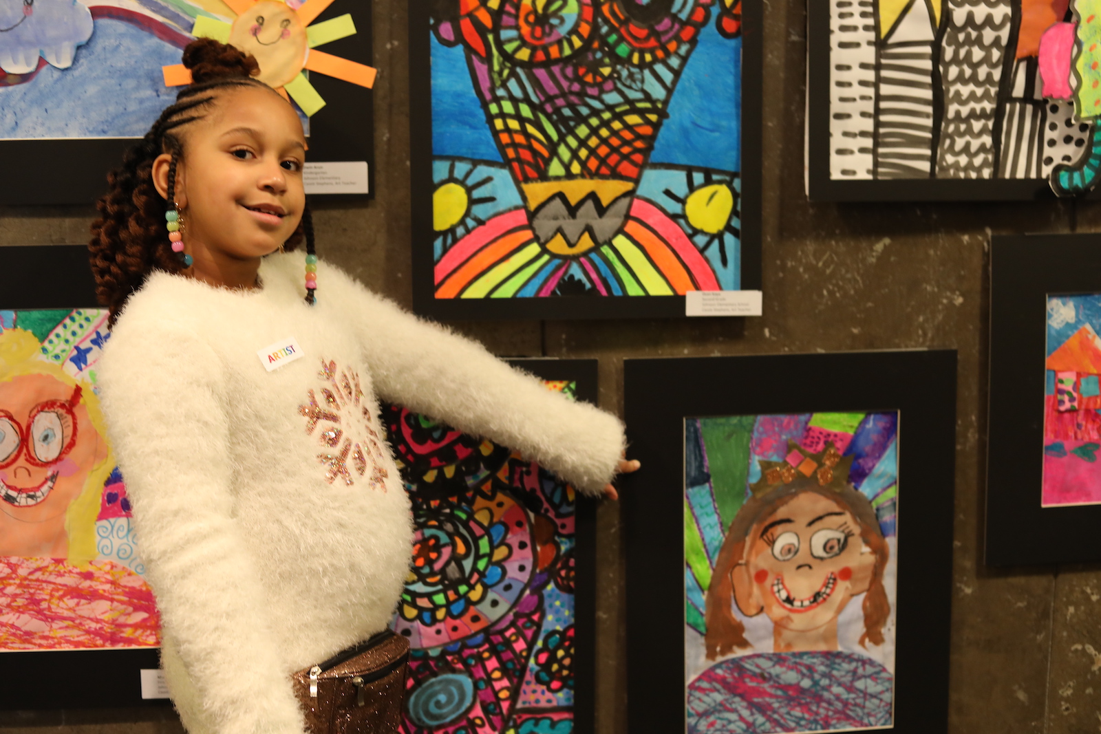 Young girl in white sweater proudly points to her artwork featured in the exhibition