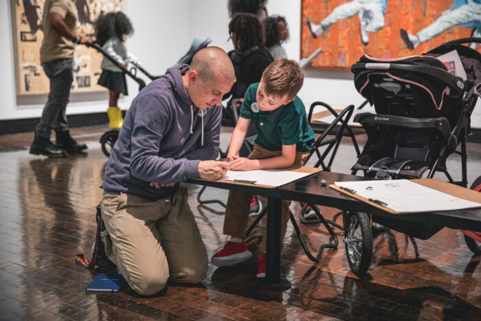 Father kneeling on ground next to son sitting on a stool as they both work on a drawing.