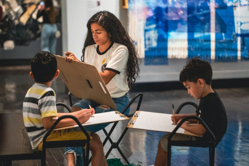 Mom and two sons sit on stools holding easels and drawing