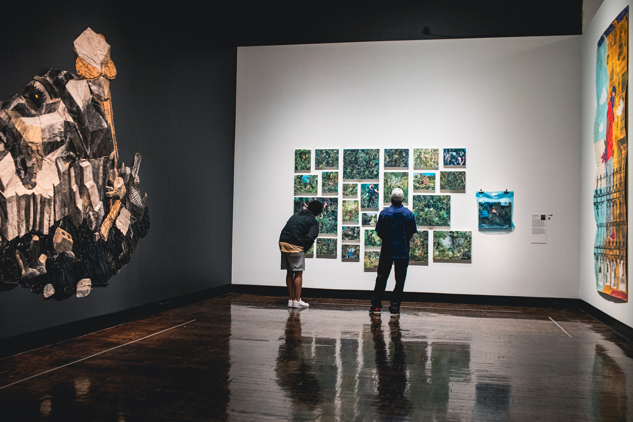 Two people looking at a multi piece artwork with two other large scale works on the walls beside them