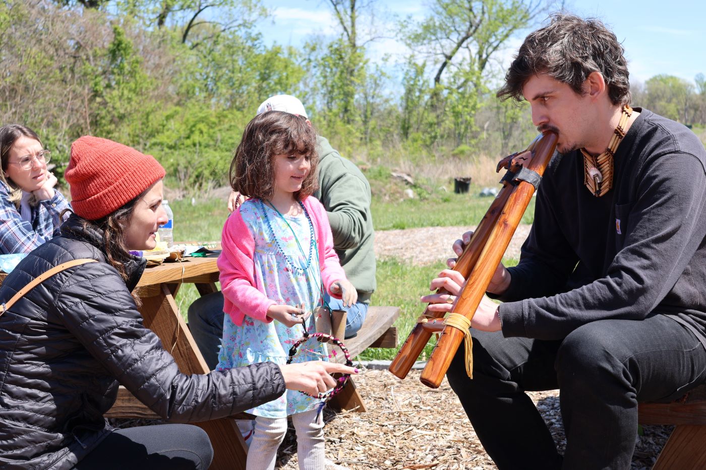 Man blowing into a wooden instrument as a mother and daughter watch