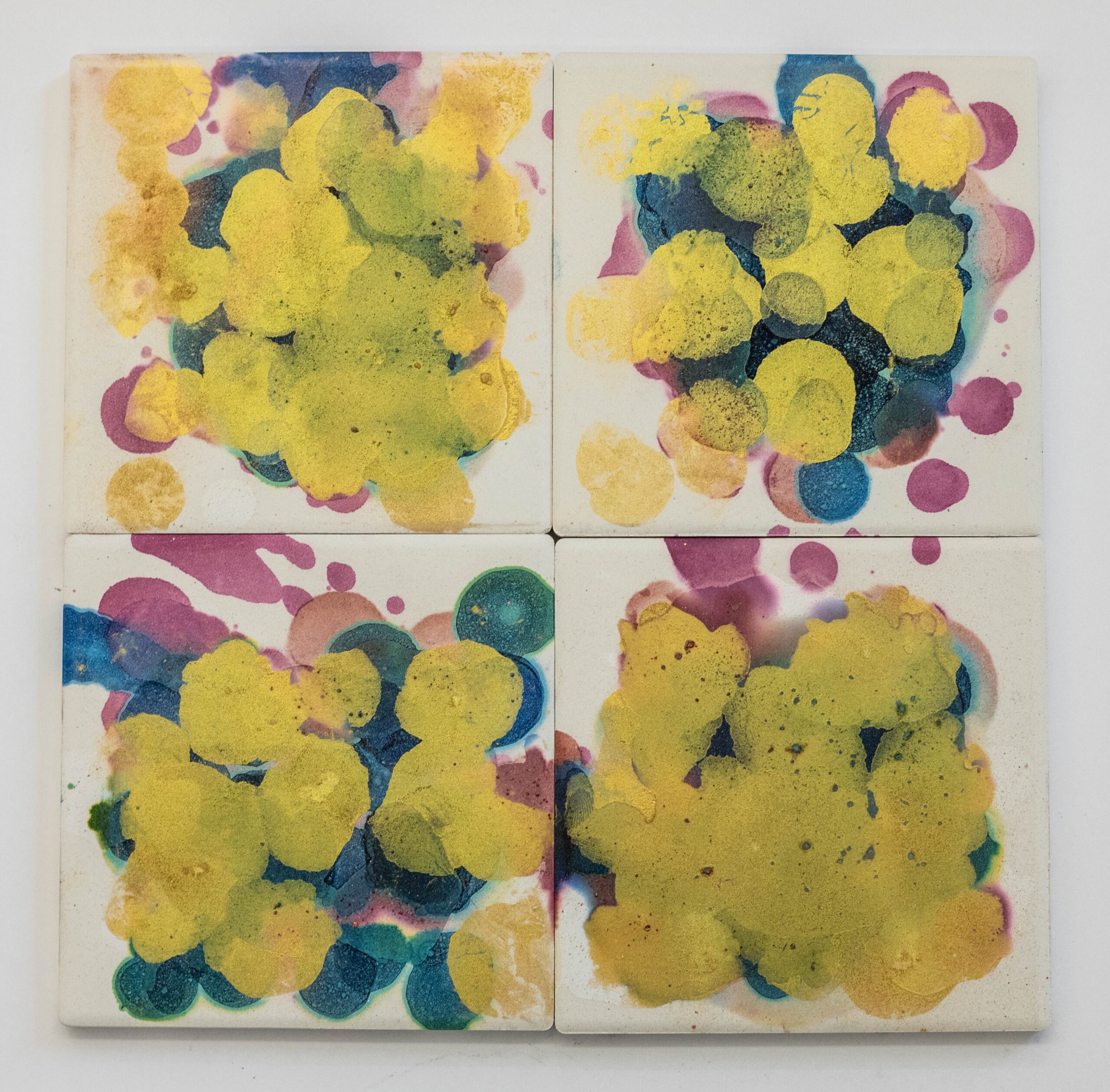 Four small tiles place together as a square and painted with circular layers of colors in yellow, blue, and magenta