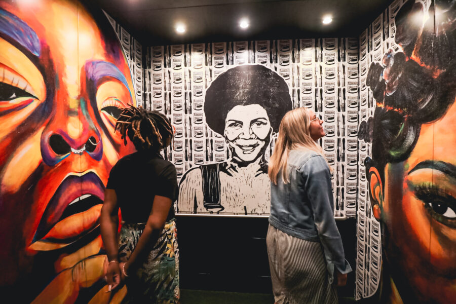 Two women standing in an elevator looking at large scale art featuring Black women applied to the wall.