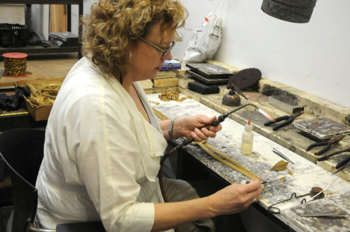 Making jewelry from metal, the artisan is in her studio, torch in hand, creating her next piece. 