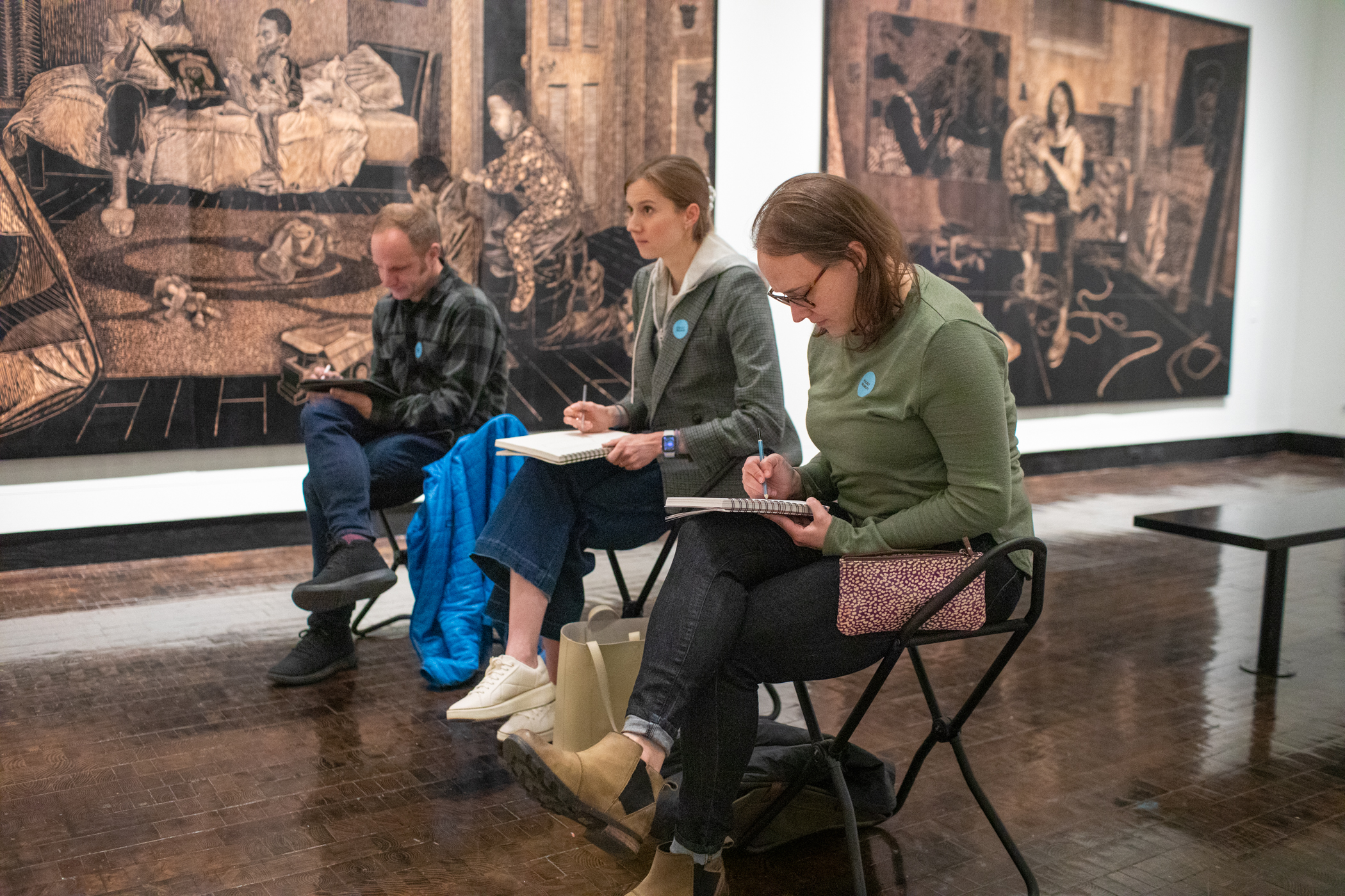 Three people sitting on stools in the galleries drawing on lap easels