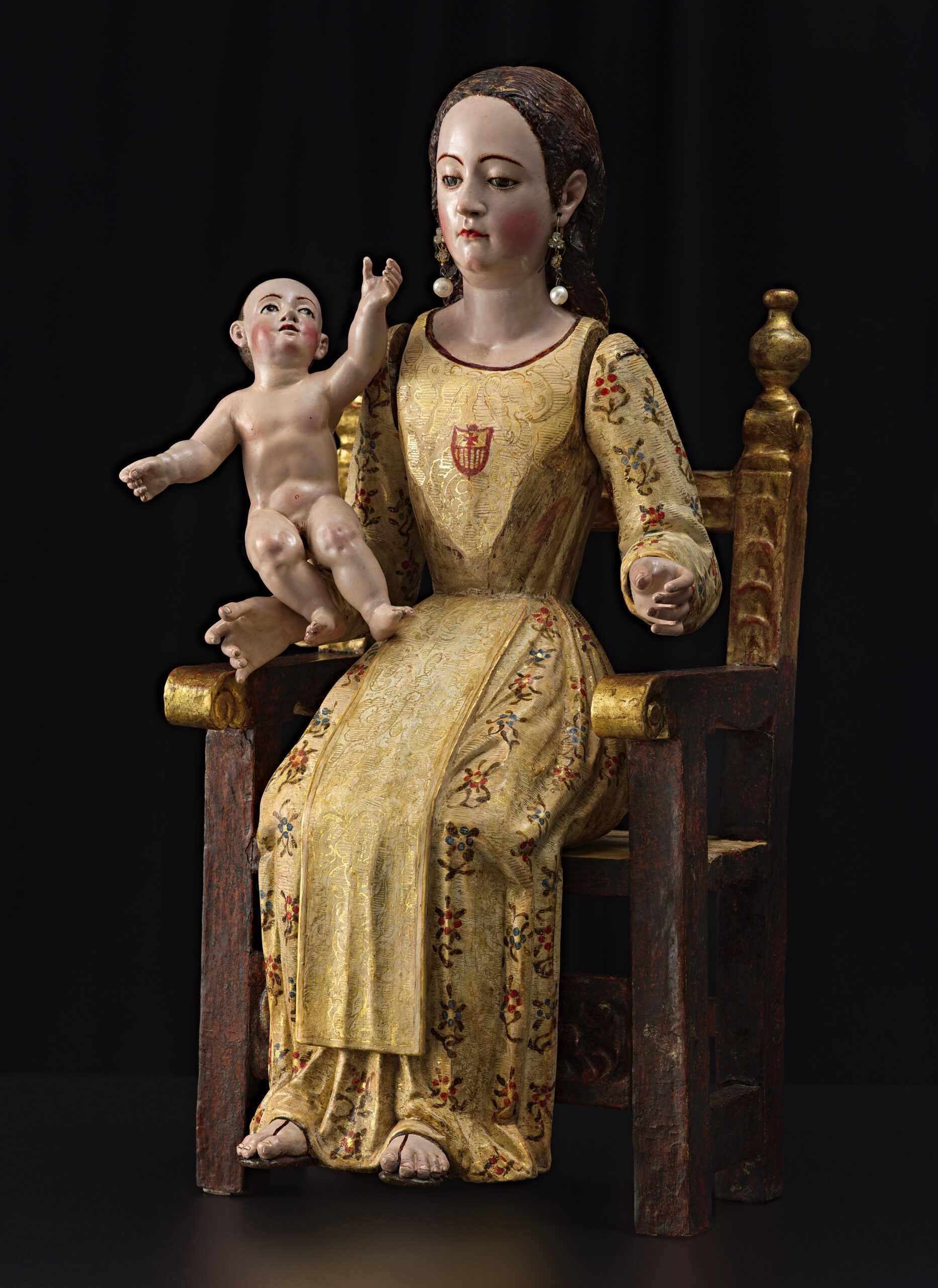 Virgin Mary and baby sitting in a wooden chair. She is wearing a long gold gown holding