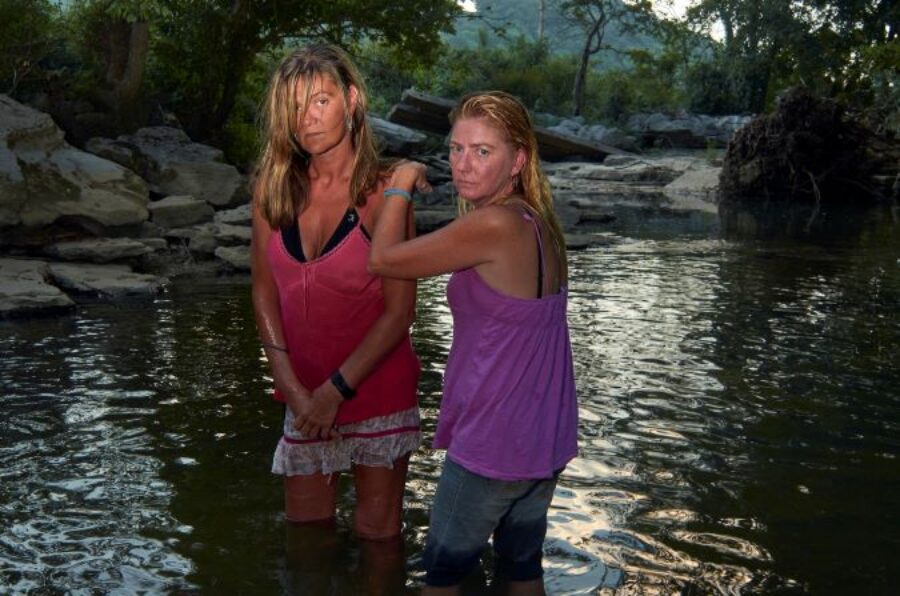 Photograph of two women with stern looks on their facing standing in the middle of a creek.