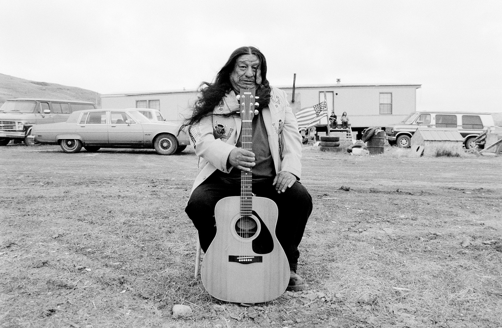 Man with long black hair sitting on a chair in front of a trailer holding a guitar.
