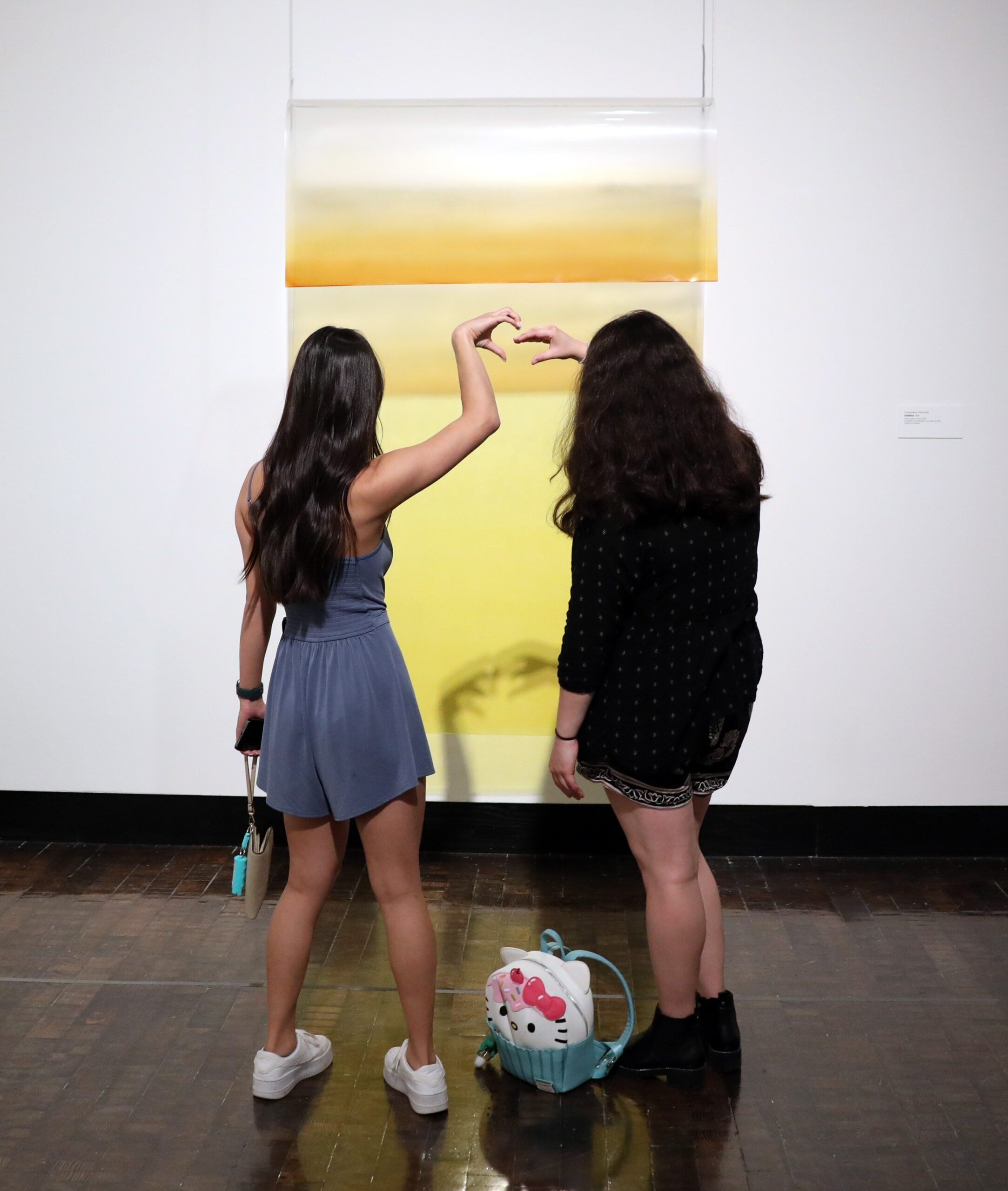 Two teens looking at a bright yellow piece of art holding up the heart symbol with their hands.