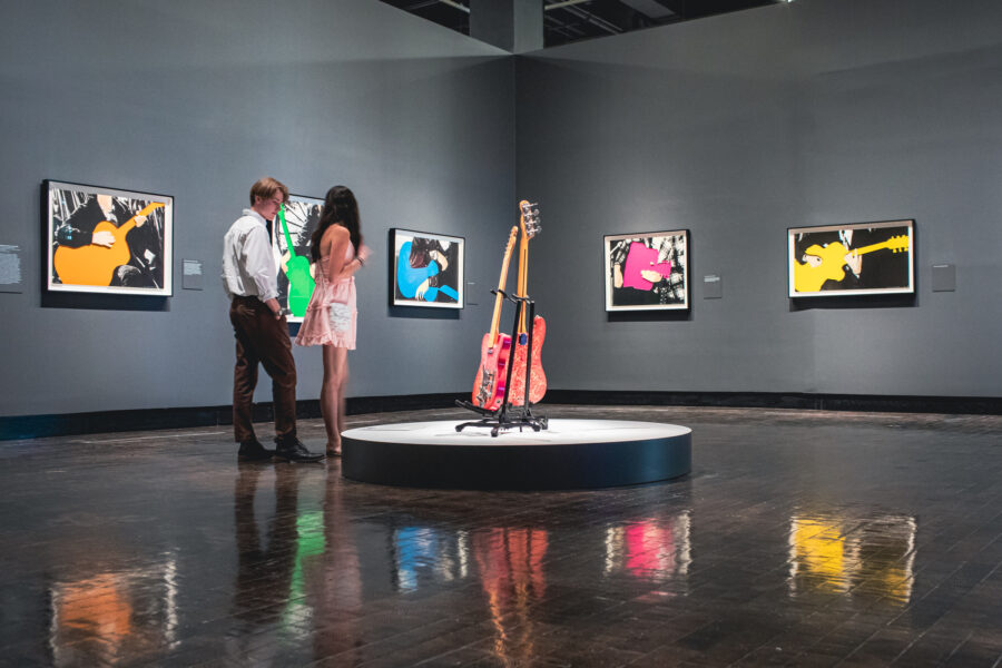 Couple walking in the galleries looking at guitar paintings and a guitar on display in the center of the room