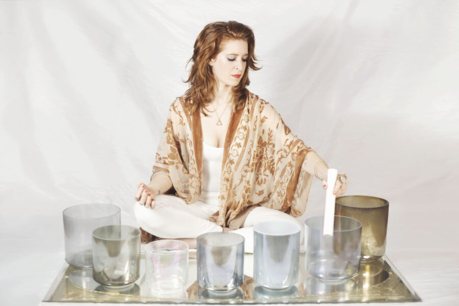 Woman sitting cross-legged surrounded by different size glass containers and holding a white rod over one of the containers